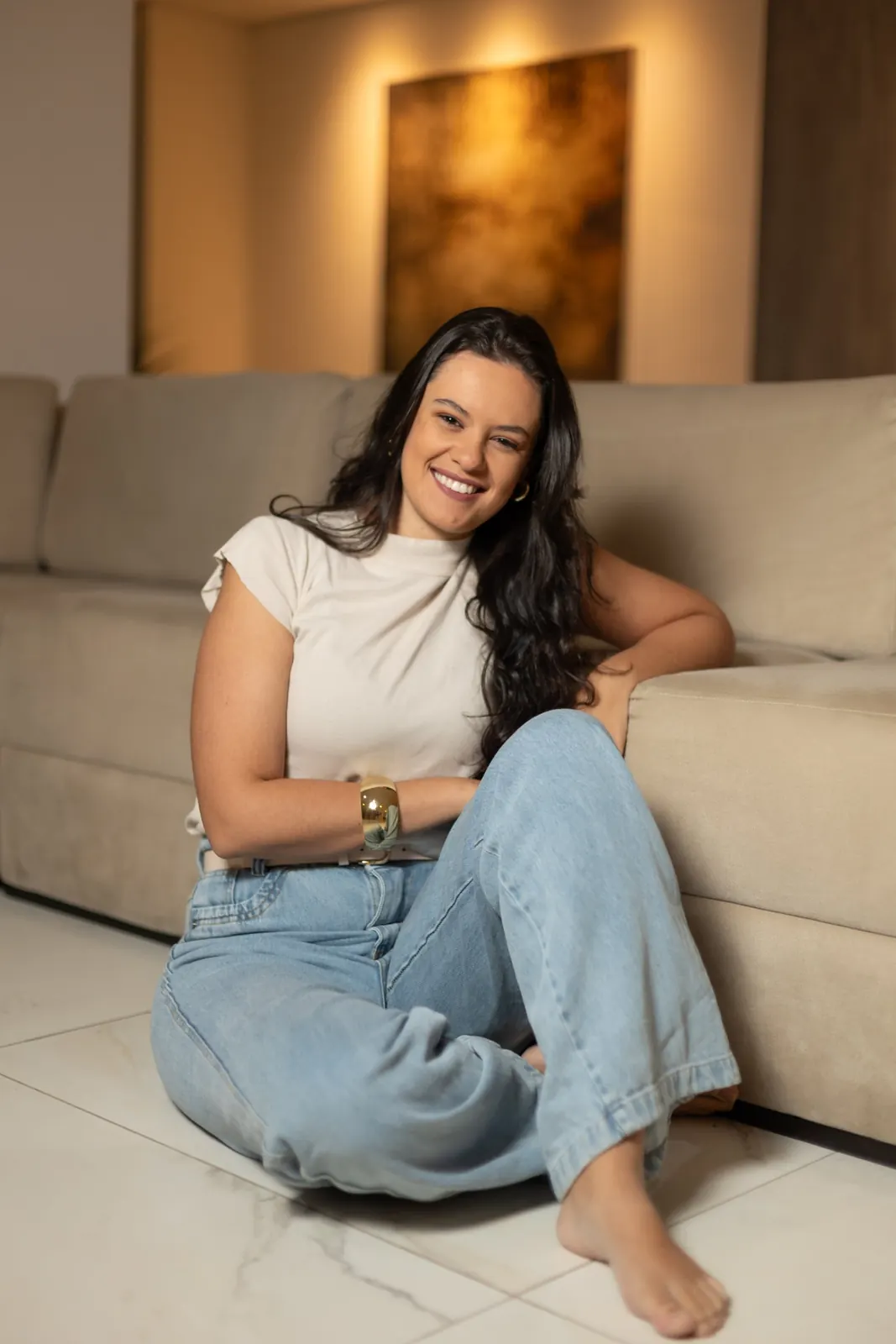 Raquel sorrindo, sentada no chão e apoiada no sofá