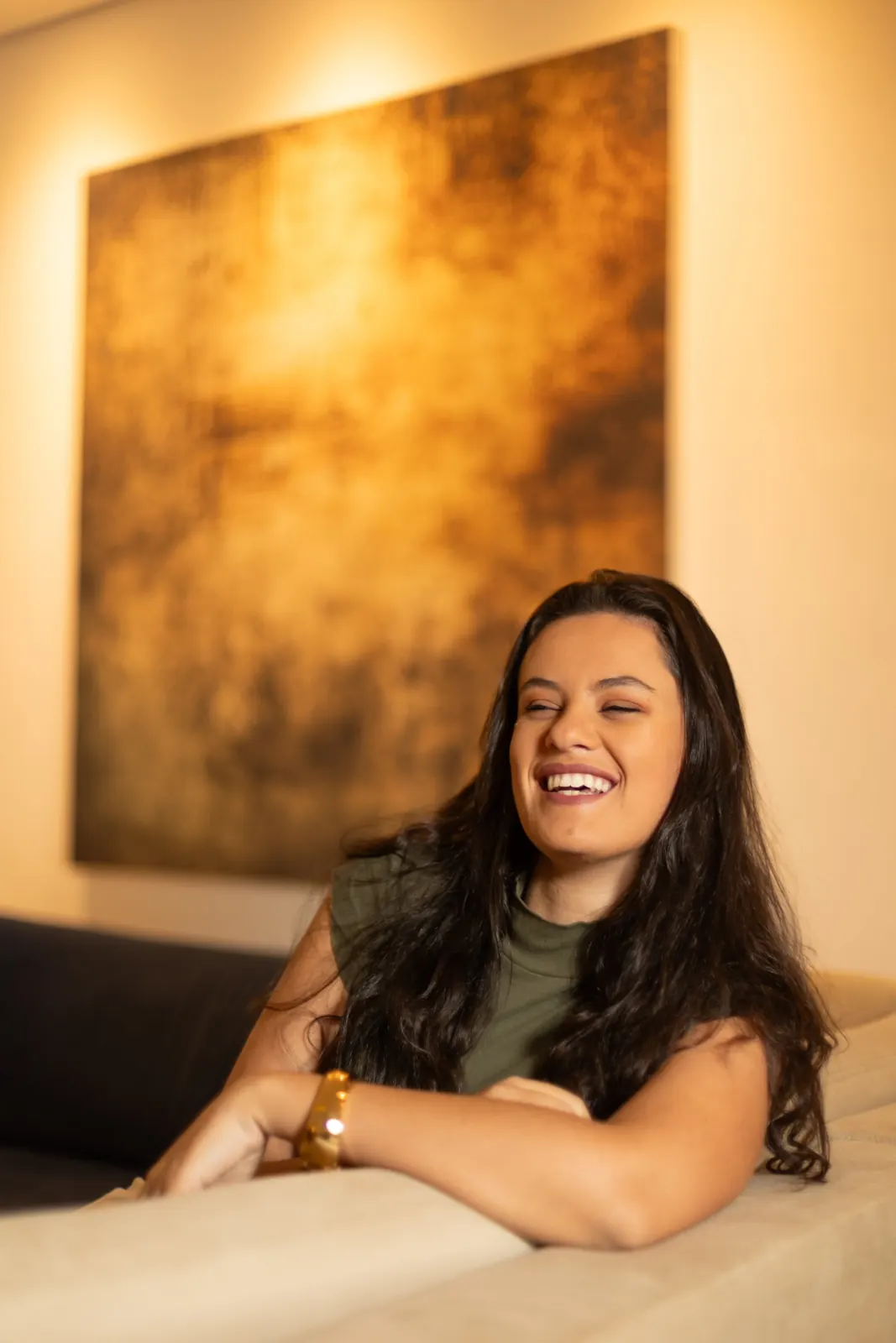 Raquel rindo, apoiada no sofá, com um quadro dourado ao fundo